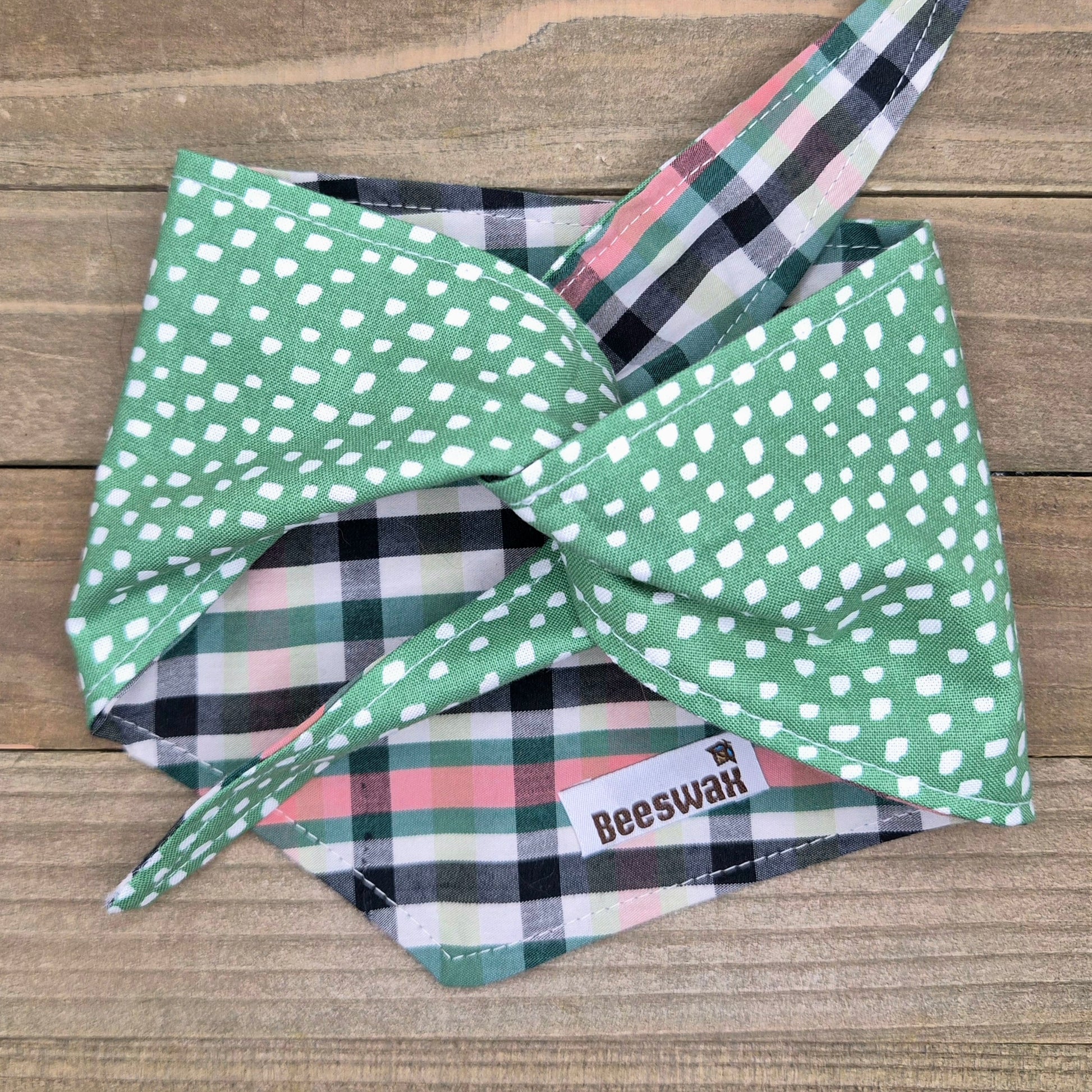A green and white polka dot bandana with a plaid pattern, tied in a bow, placed on a wooden surface.