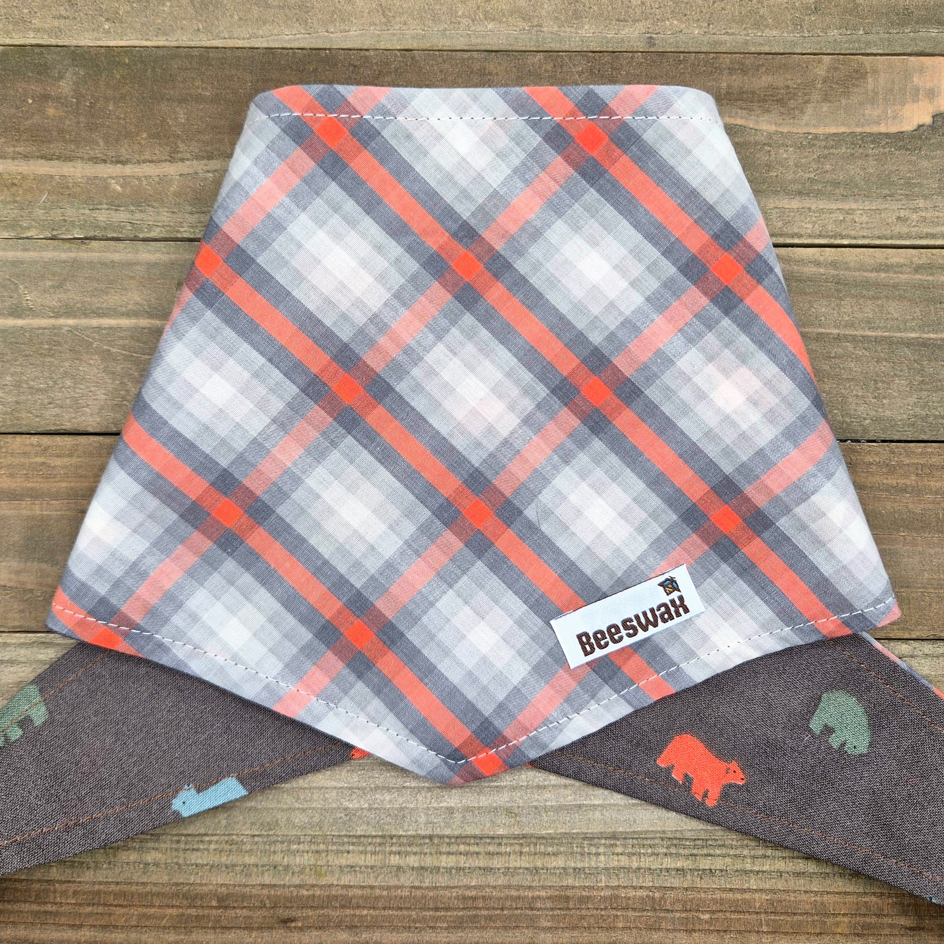 A plaid dog bandana with a gray and orange pattern, featuring a small bear design, is displayed on a wooden surface.