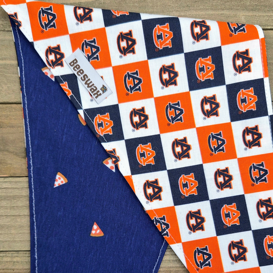 a dog bandana with a pattern of orange and white squares, each containing a small orange triangle. The bandana is placed on a wooden surface, and there is a tag attached to it.