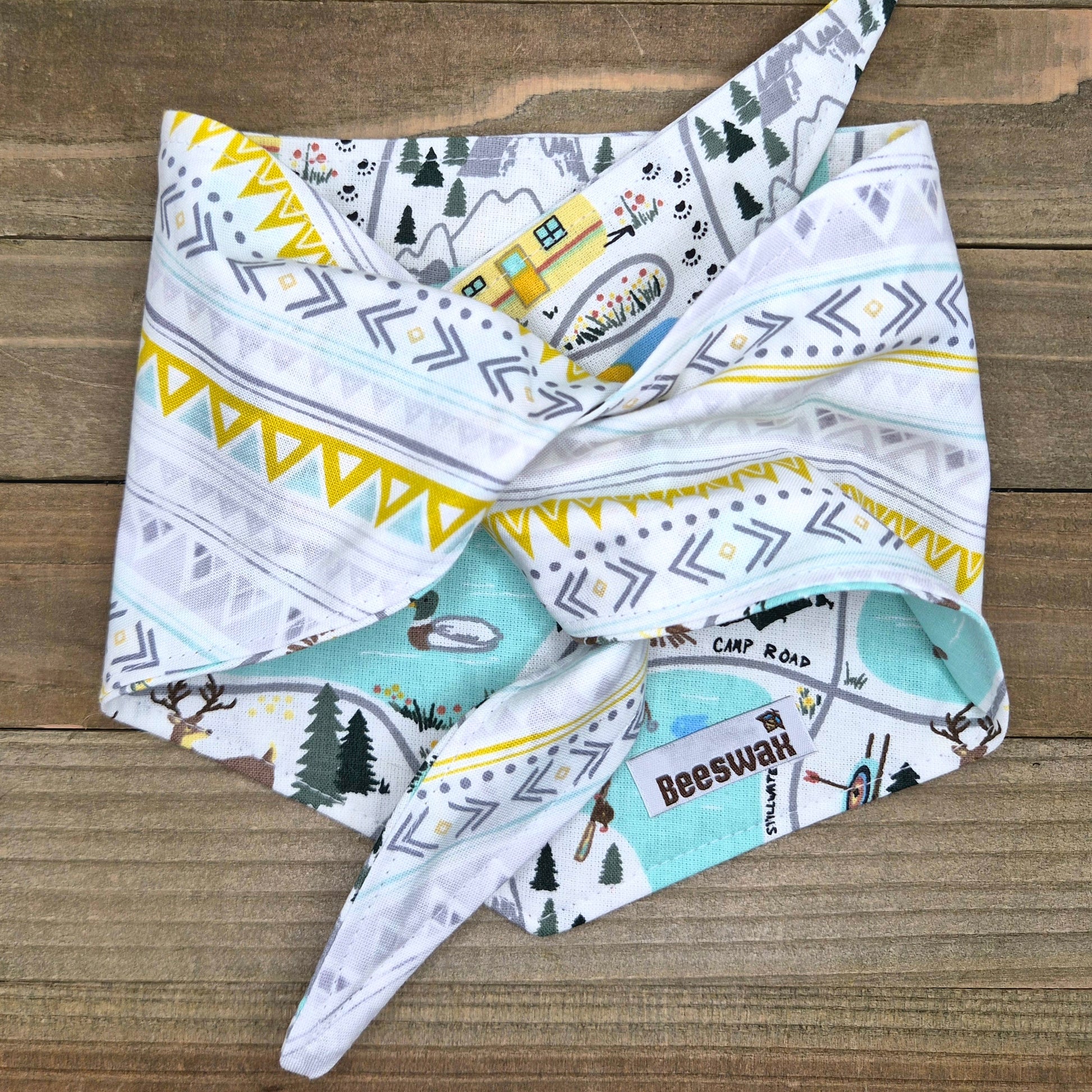 A colorful bandana with a pattern of trees, mountains, and a house, folded and placed on a wooden surface.