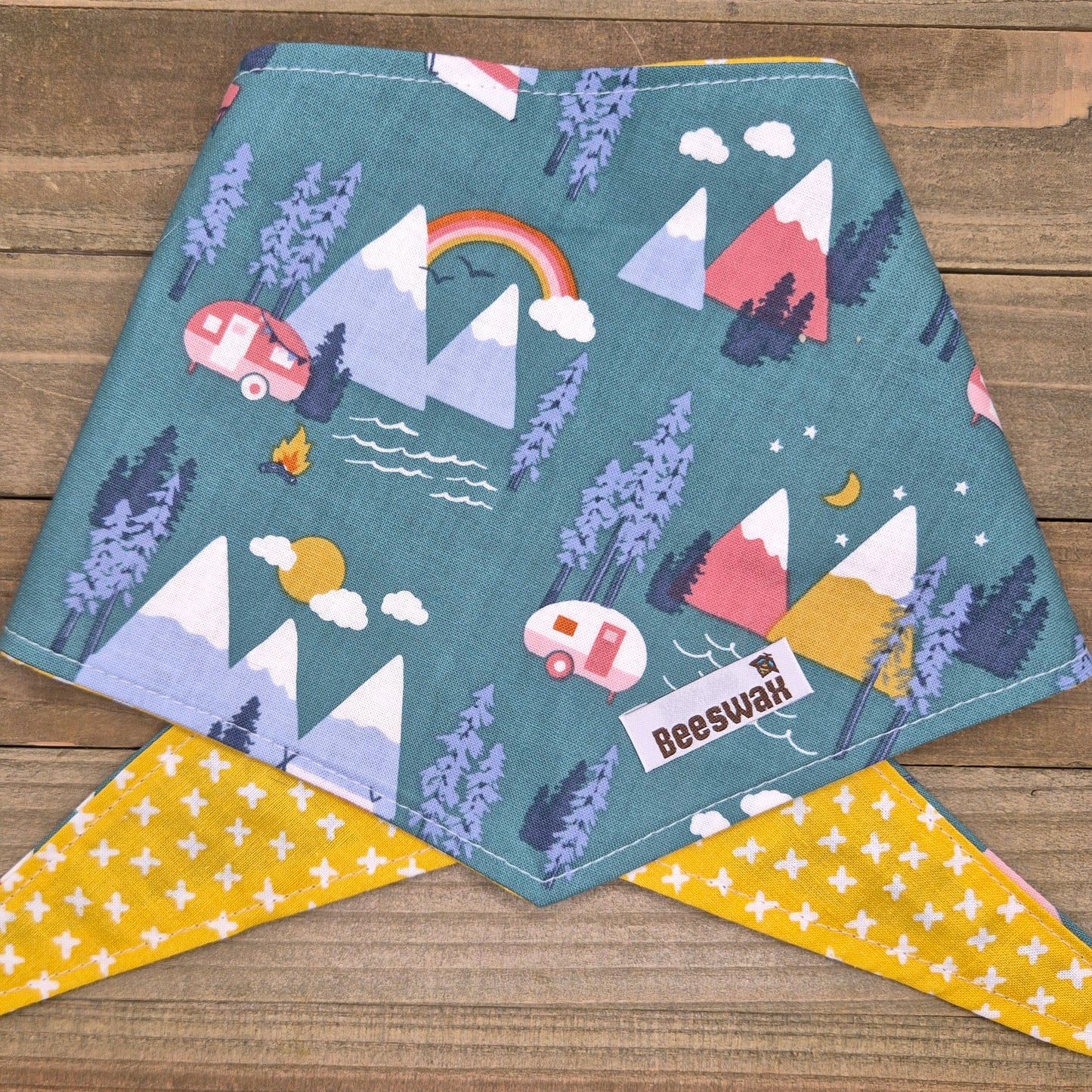A colorful bandana with a mountain and camping scene printed on it, featuring a rainbow, camper van, pine trees, and other natural elements.