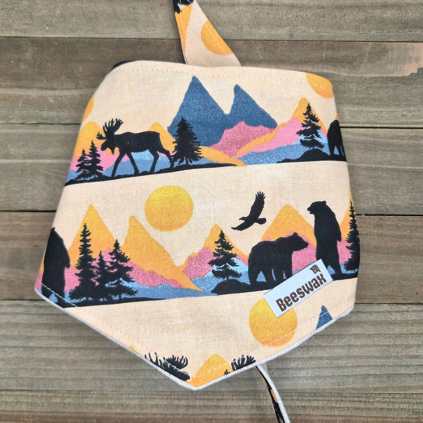 A beige fabric bandana with a printed design depicting silhouettes of animals, including a moose, a bear, and a bird, against a mountain landscape with a sun.