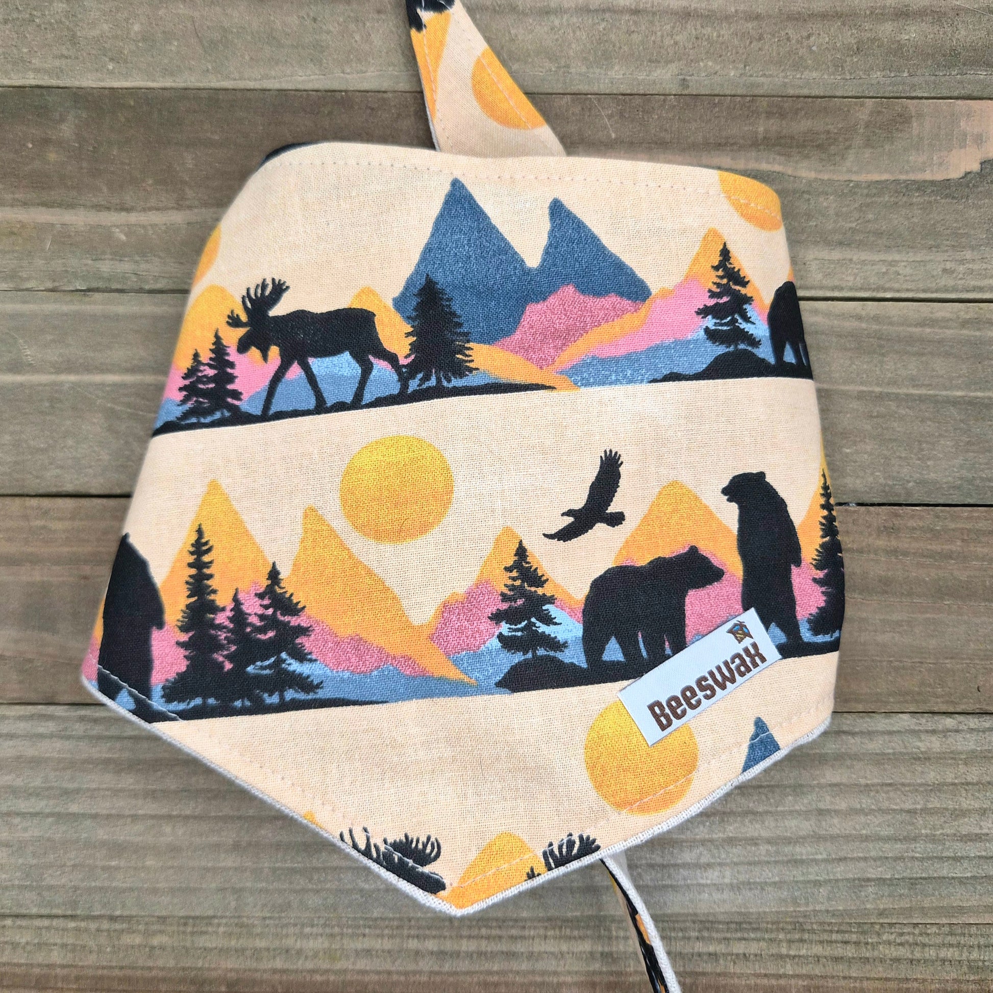 A beige fabric bandana with a printed design depicting silhouettes of animals, including a moose, a bear, and a bird, against a mountain landscape with a sun.